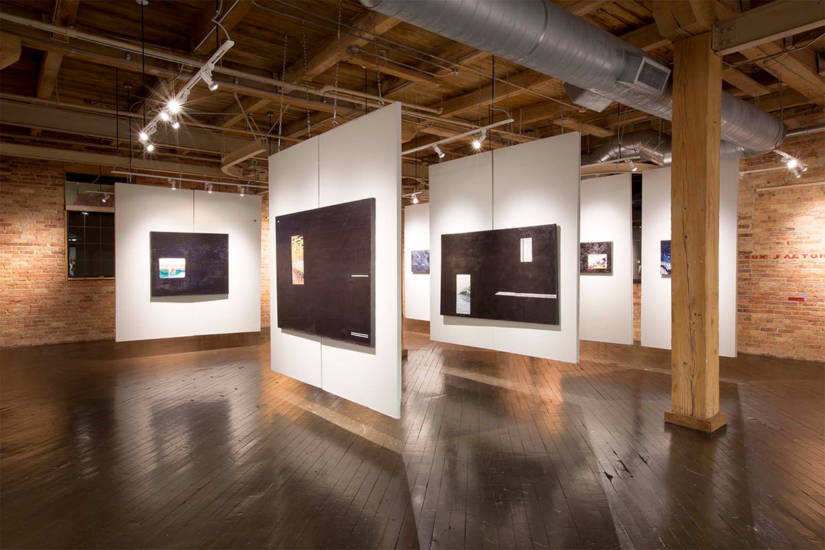 Visitors explore an art gallery with brick walls and wooden floors, featuring paintings, photographs, and sculptures at the Box Factory for the Arts.