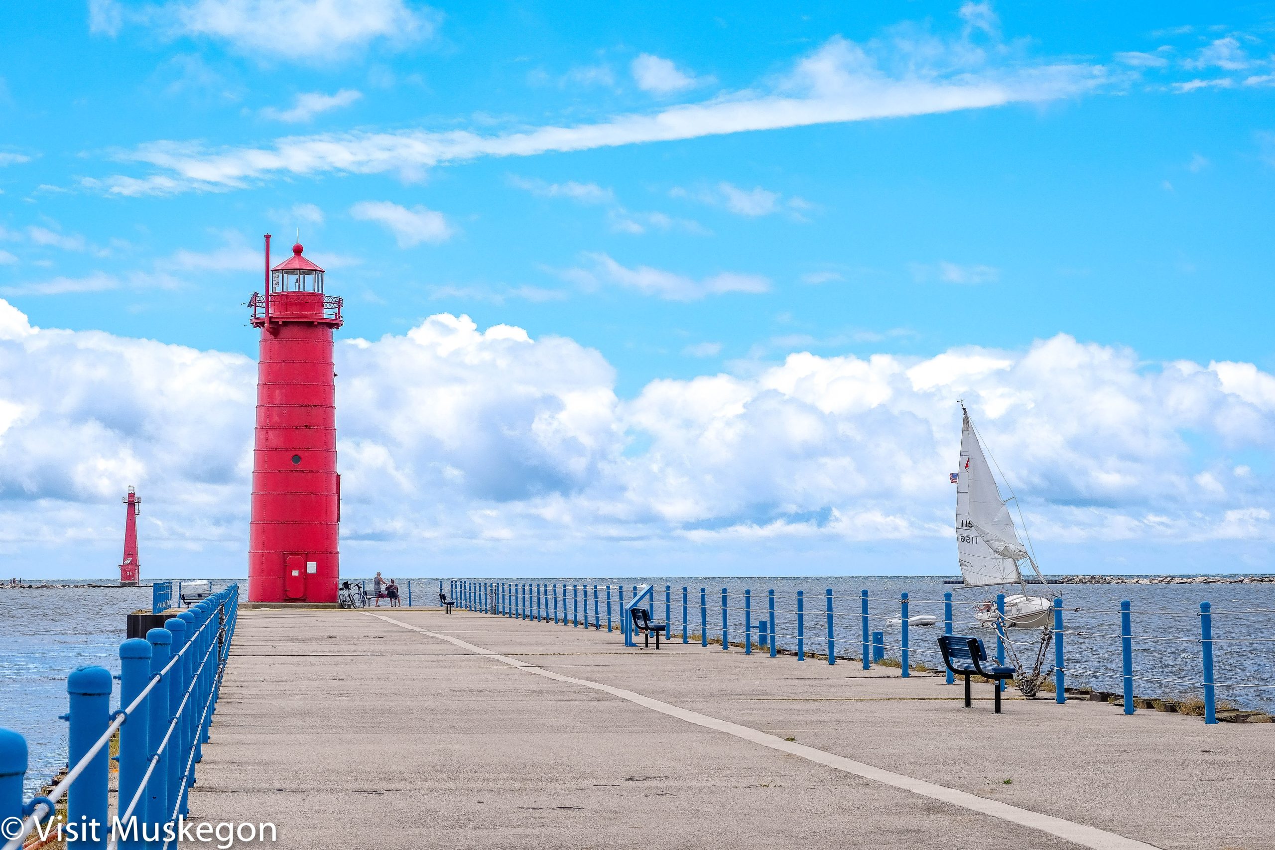Two red light towers stand in front of blue sky filled with cumulus clouds. a sailboat with white sails is seen to the right of pier that leads to one of the lighttowers.