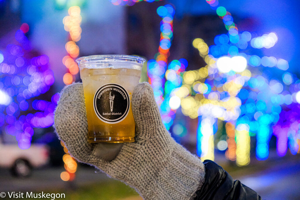 mitten covered hand holds beverage up against glowing holiday lights