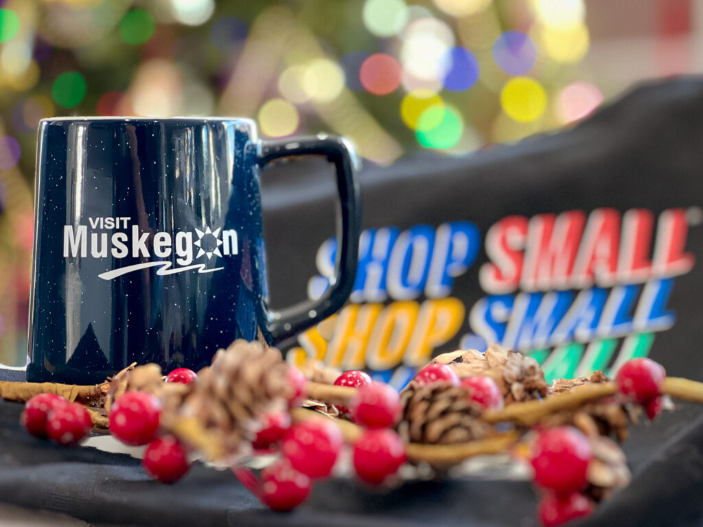blue mug with text that reads "visit muskegon" sits on "shop small" bag among small pines and berries. holiday lights glow in background. 