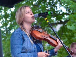 young blonde woman with side braid and denim jacket sings into microphone and plays violin in park