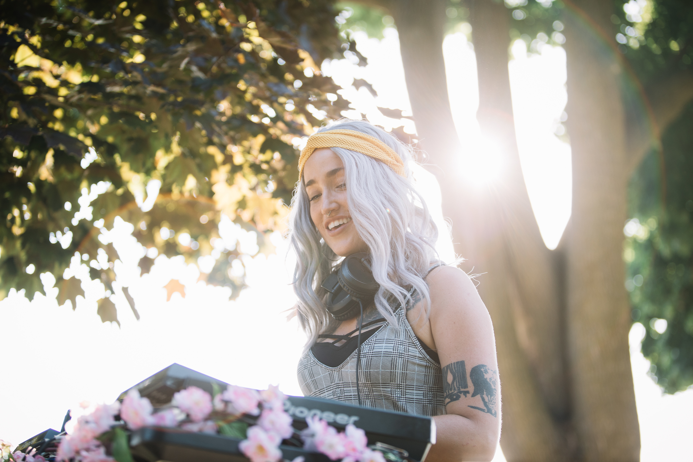 young female deejay with long hair in yellow headband stands at music board in sunlit park filled with trees and light.