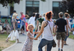 mom and daughter dance on wide sidewalk in park surround by people