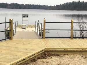 View of wooden dock leading to an accessible kayak launch, extending into a calm lake, surrounded by tranquil forest scenery.