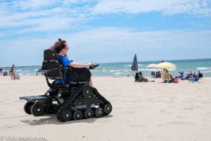 A person using a tracked wheelchair on a sandy beach, looking towards Lake Michigan where people are enjoying the sunny day under umbrellas. The scene includes a clear blue sky and gentle waves