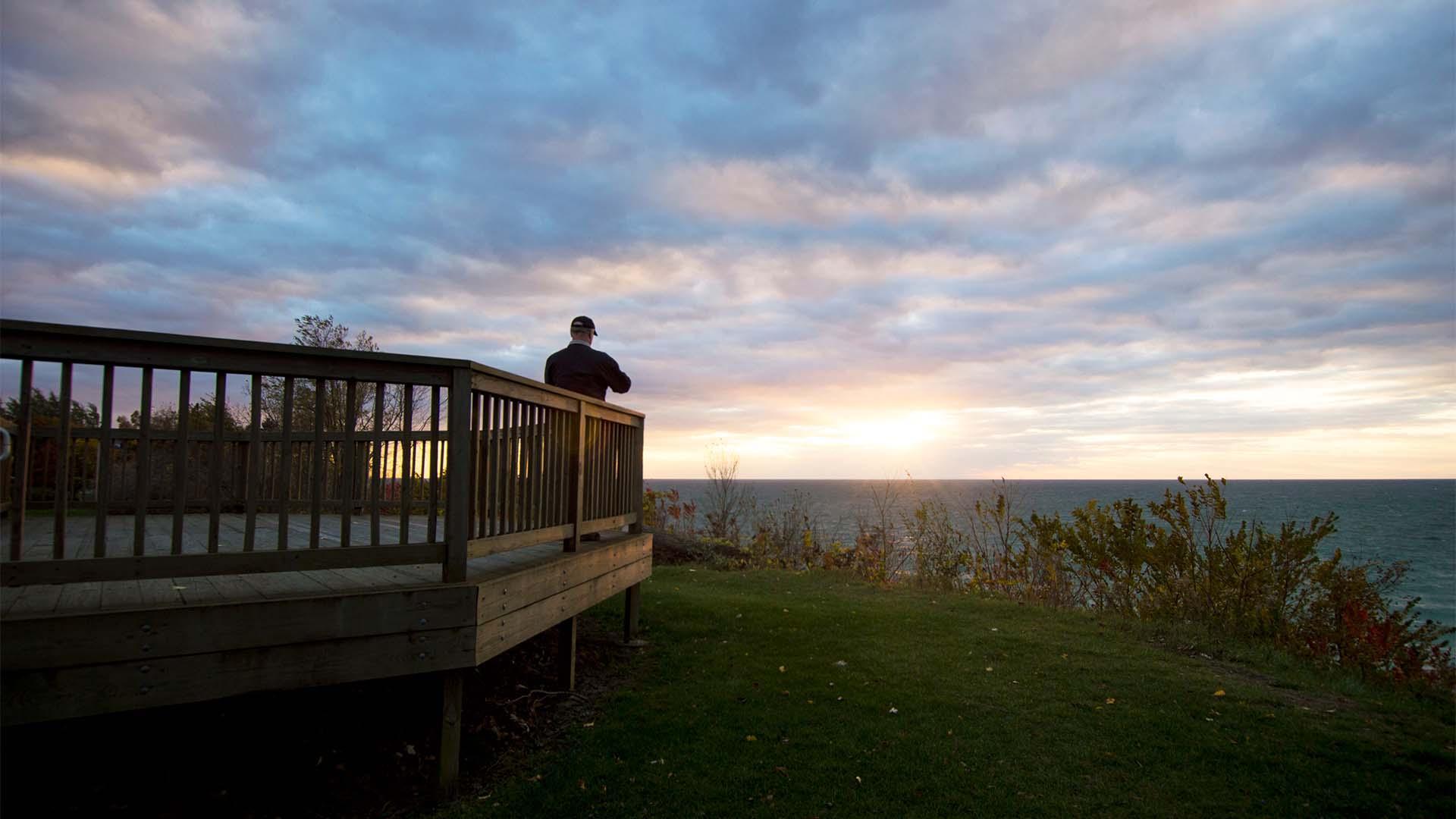 A person stands on a wooden deck overlooking Lake Michigan as the sun sets, casting warm colors over the water and sky at Lookout Park.