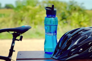 Bicycle seat, blue water bottle, and black helmet rest on a wooden table outdoors, with blurred greenery in the background, suggesting a casual biking outing.