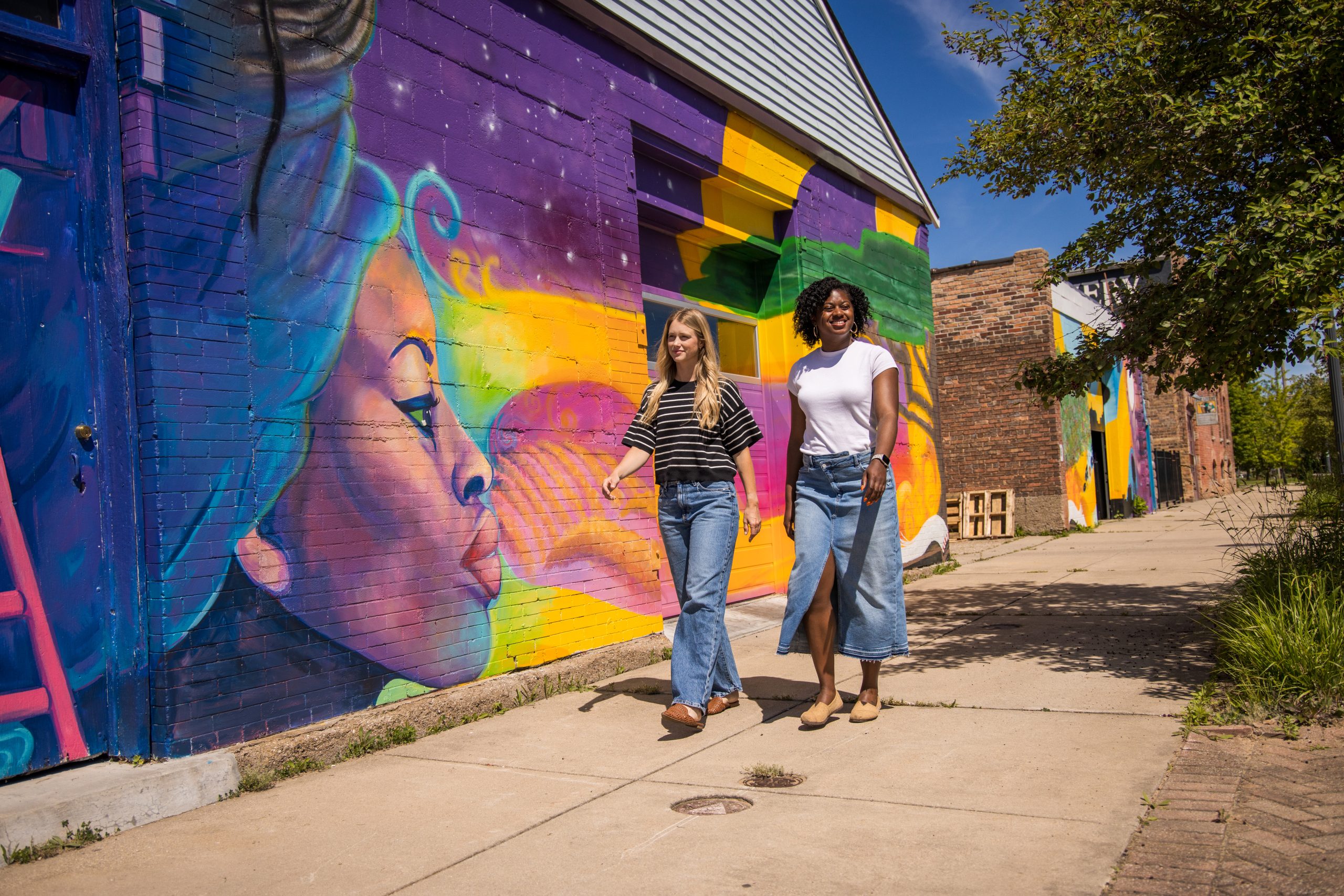 Two friends stroll past a vibrant mural of a woman in the Benton Harbor Arts District.