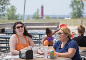 Two women sit laughing at a beachside picnic table, with a red lighthouse in the background. Bright and sunny atmosphere, relaxed and cheerful mood.