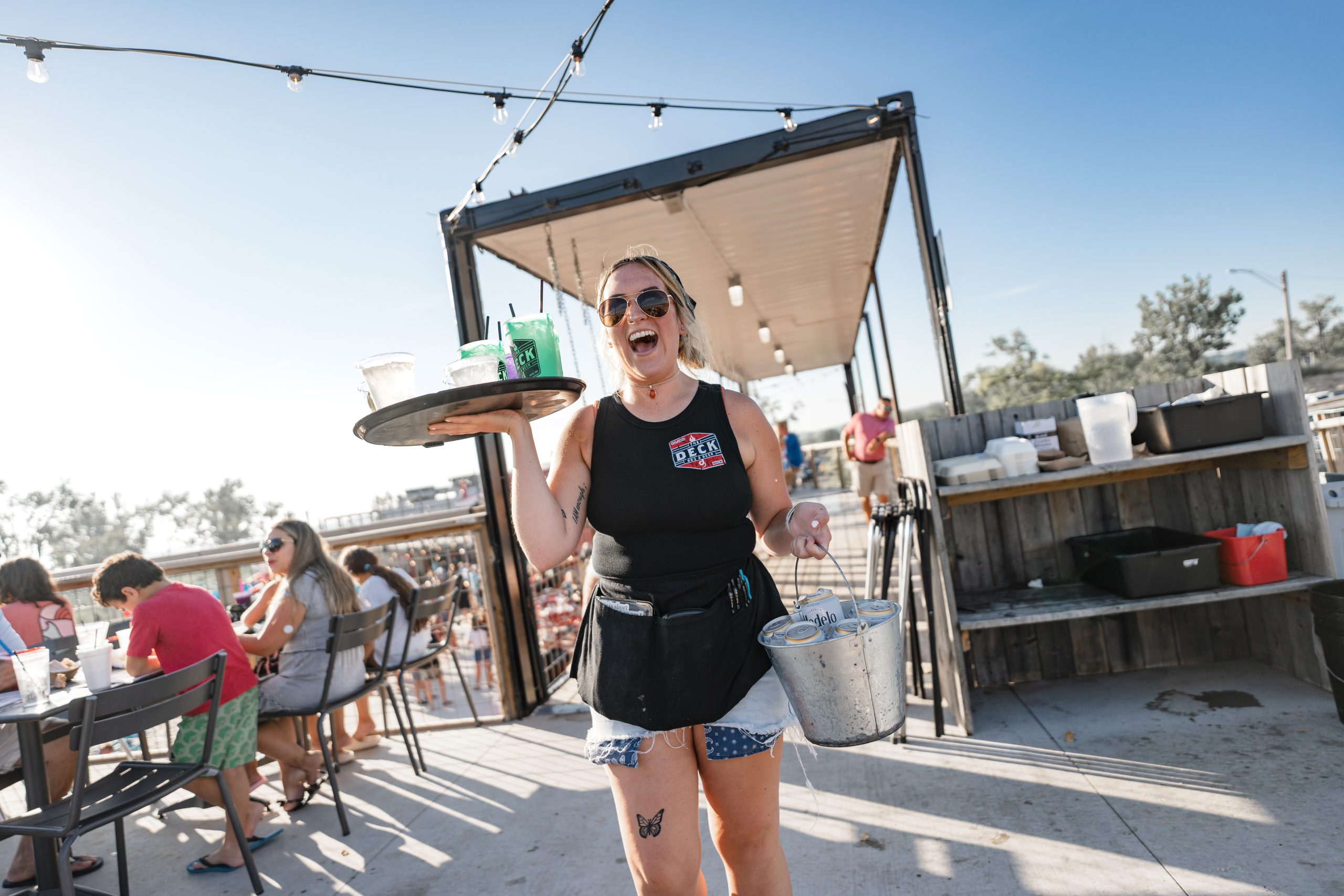 Smiling waitress with sunglasses holds a tray and bucket outdoors. Diners sit at tables under string lights in a sunny, casual setting.